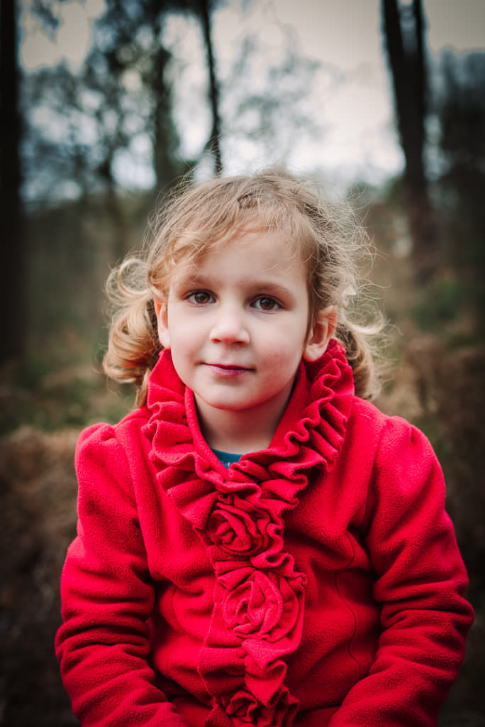 girl in red coat sits to have her photo taken on a woodland photo shoot