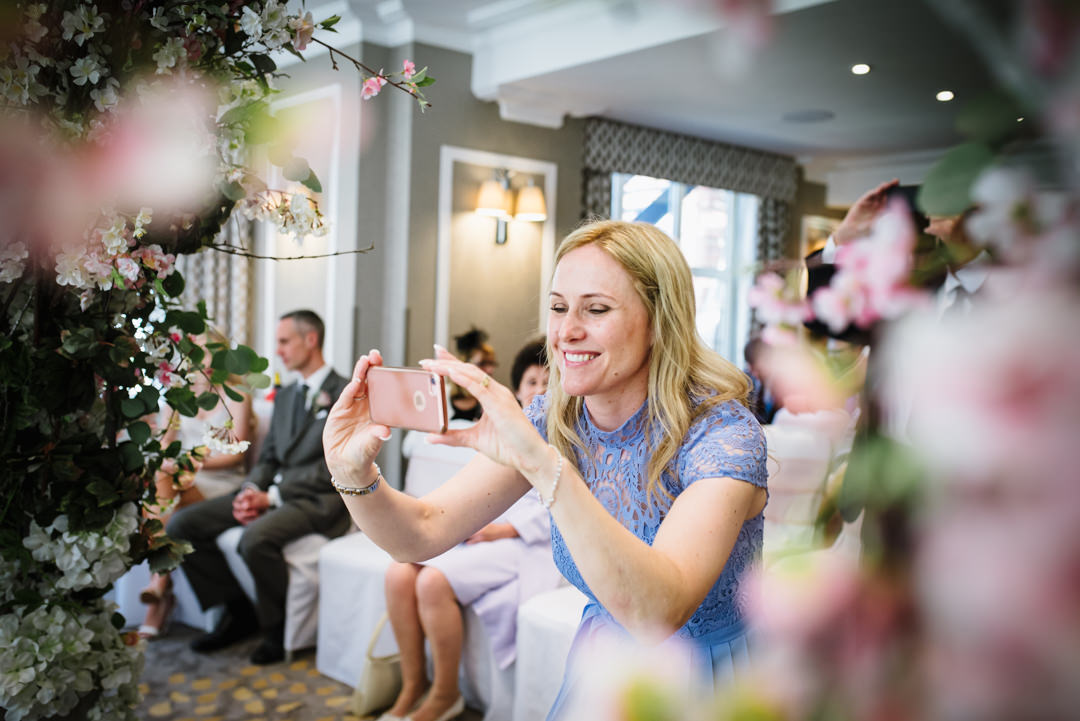 pretty female guest takes a photo of the newly weds framed by flower arch gerrards cross wedding