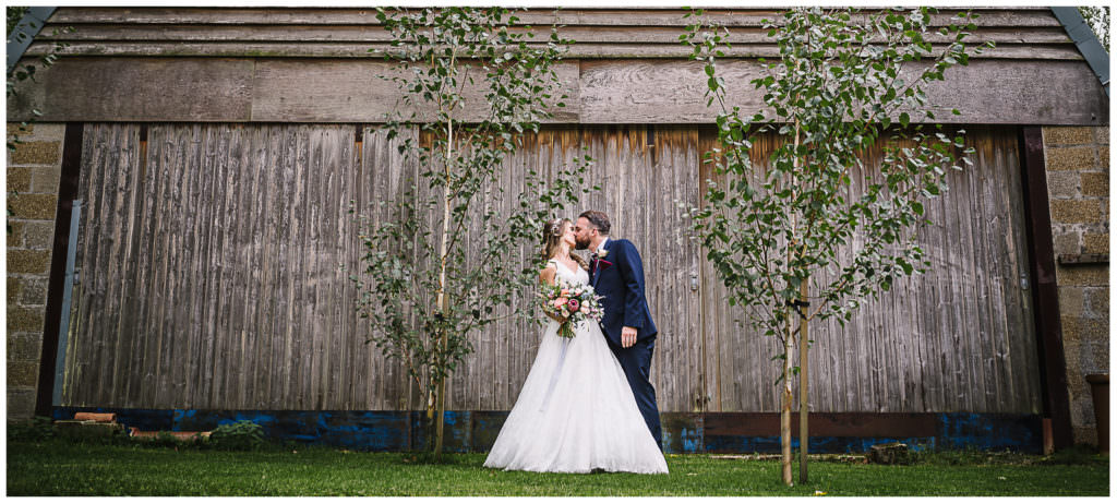 bride and groom kiss in front of the farm buildings at houchins farm small wedding venue