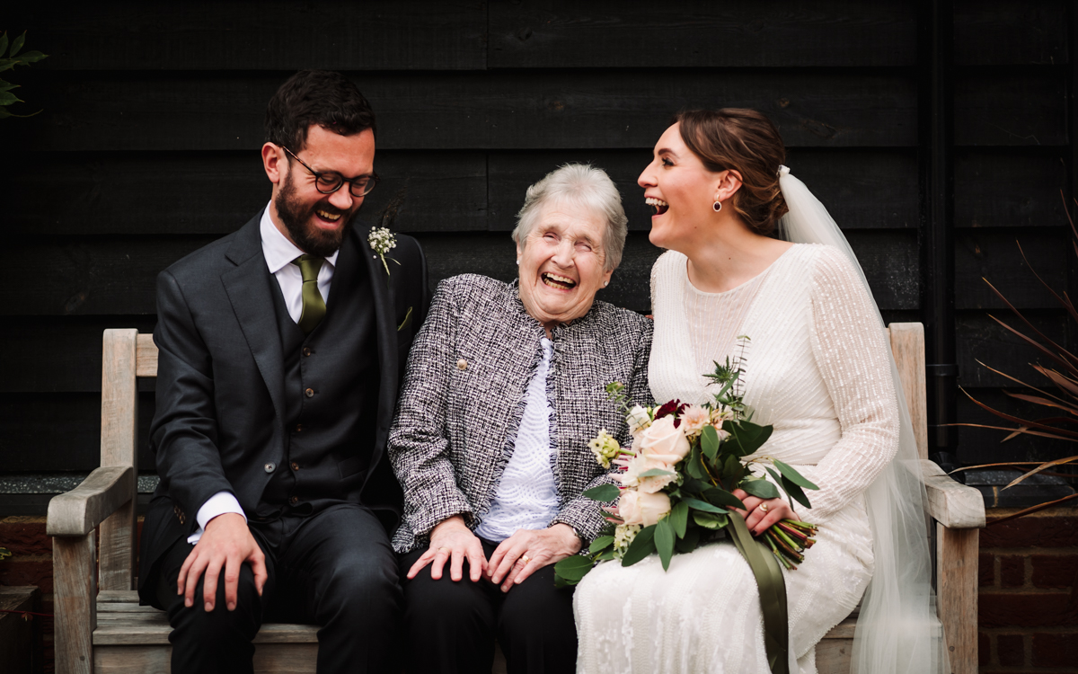 Newly weds and their grandmother sit on a bench laughing at redcoats Farm in Hertfordshire