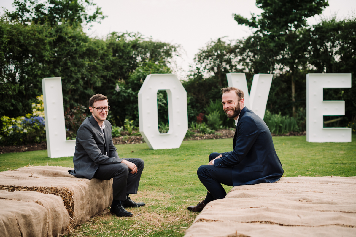 Two male guests sit on straw bales in front of love letters.