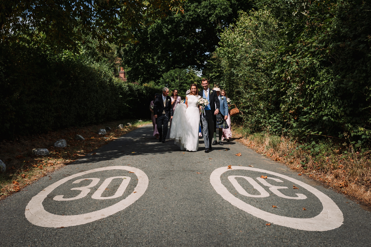 Bride and groom and their guests walk to the farmhouse where their festival style wedding is being held